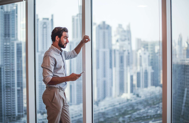 Professional looking out of a skyscraper window with more skyscrapers in the background. He has a mobile phone in his hand.