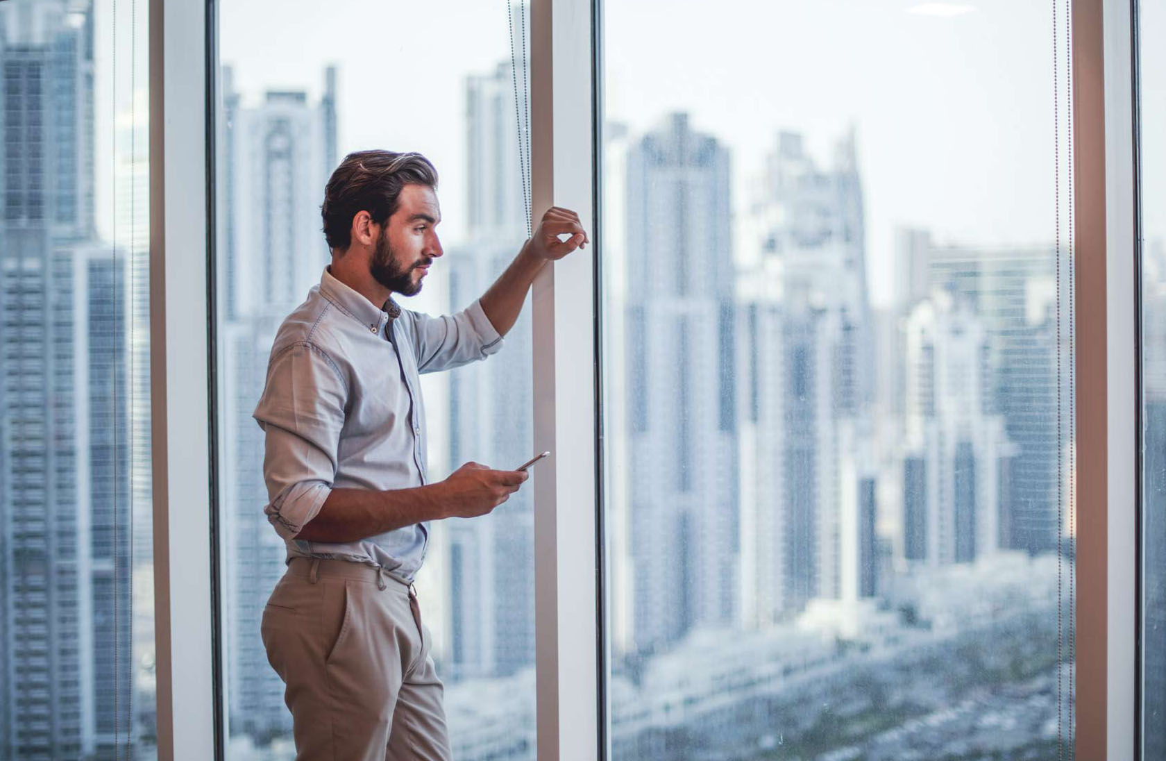 Professional looking out of a skyscraper window with more skyscrapers in the background. He has a mobile phone in his hand.