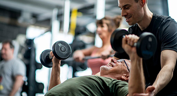 A man assists another man who is older than him as he lifts weights in the gym